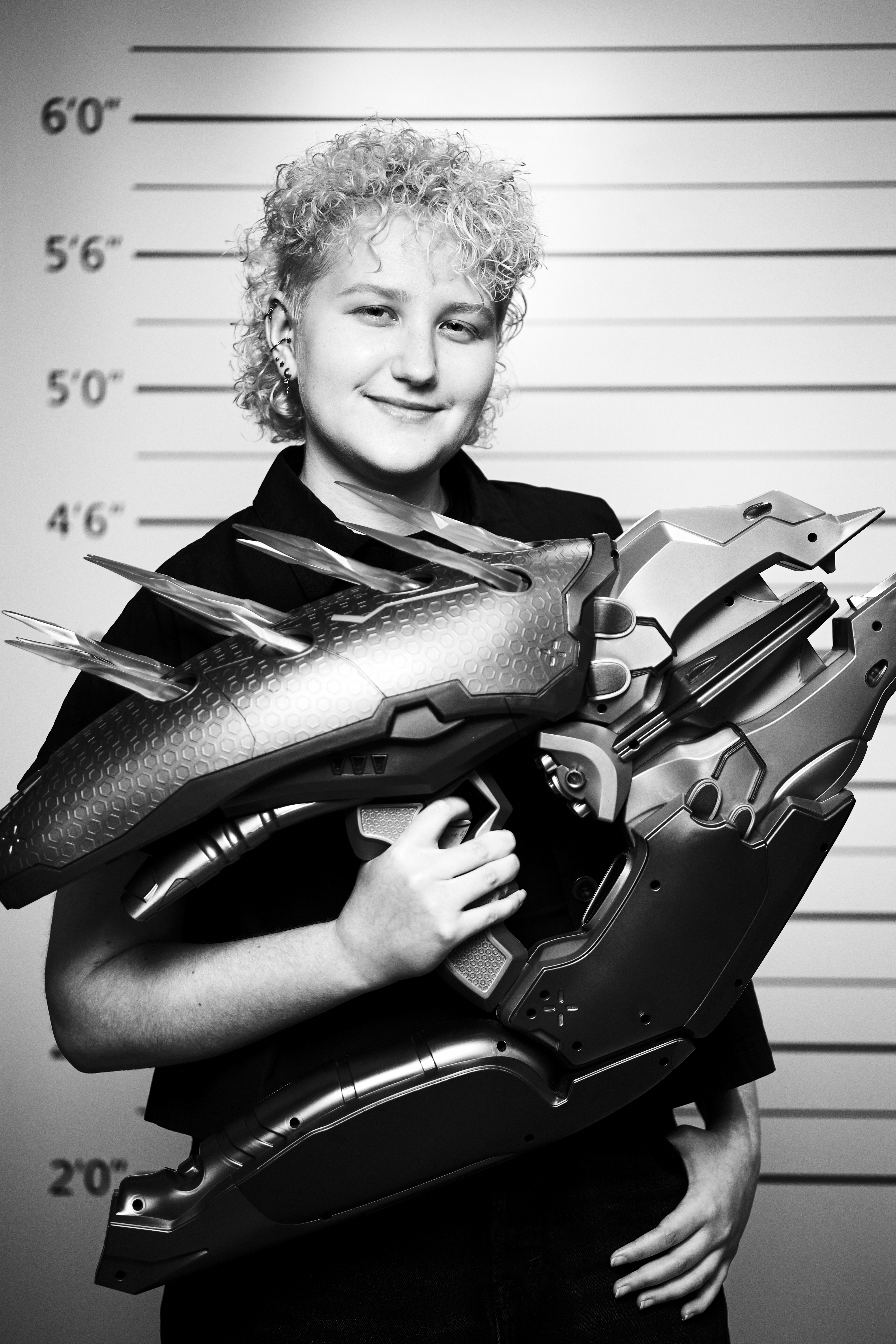 Hanna Harmon standing in front of a height measurement backdrop, smiling and holding a large decorative weapon prop